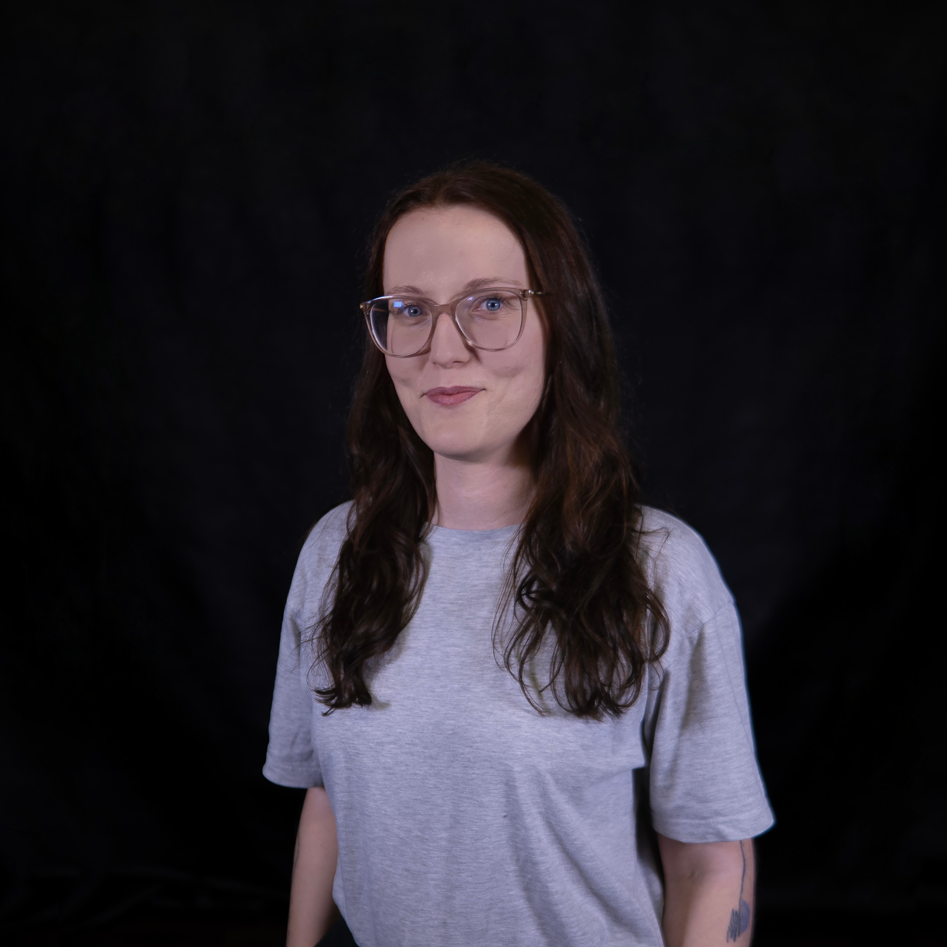 a photo of a dark-haired woman with glasses, wearing a gray t-shirt, with abstract tattoos on arms